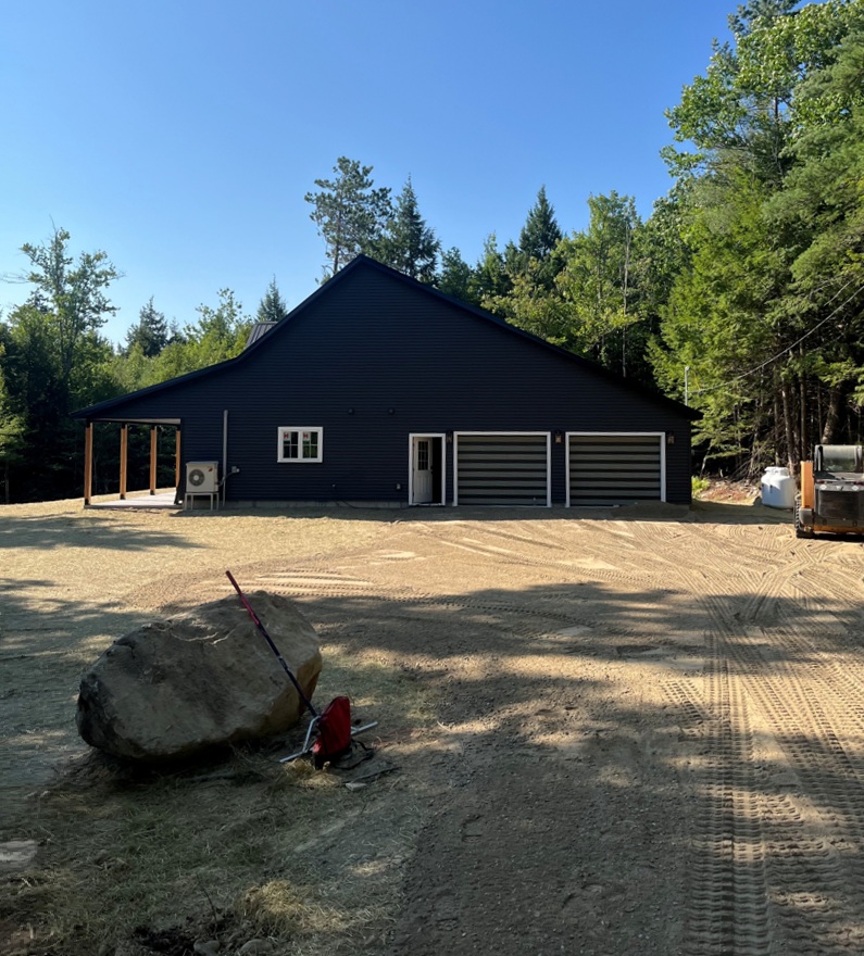 A freshly graded gravel driveway in front of a dark garage on a wooded Maine property.
